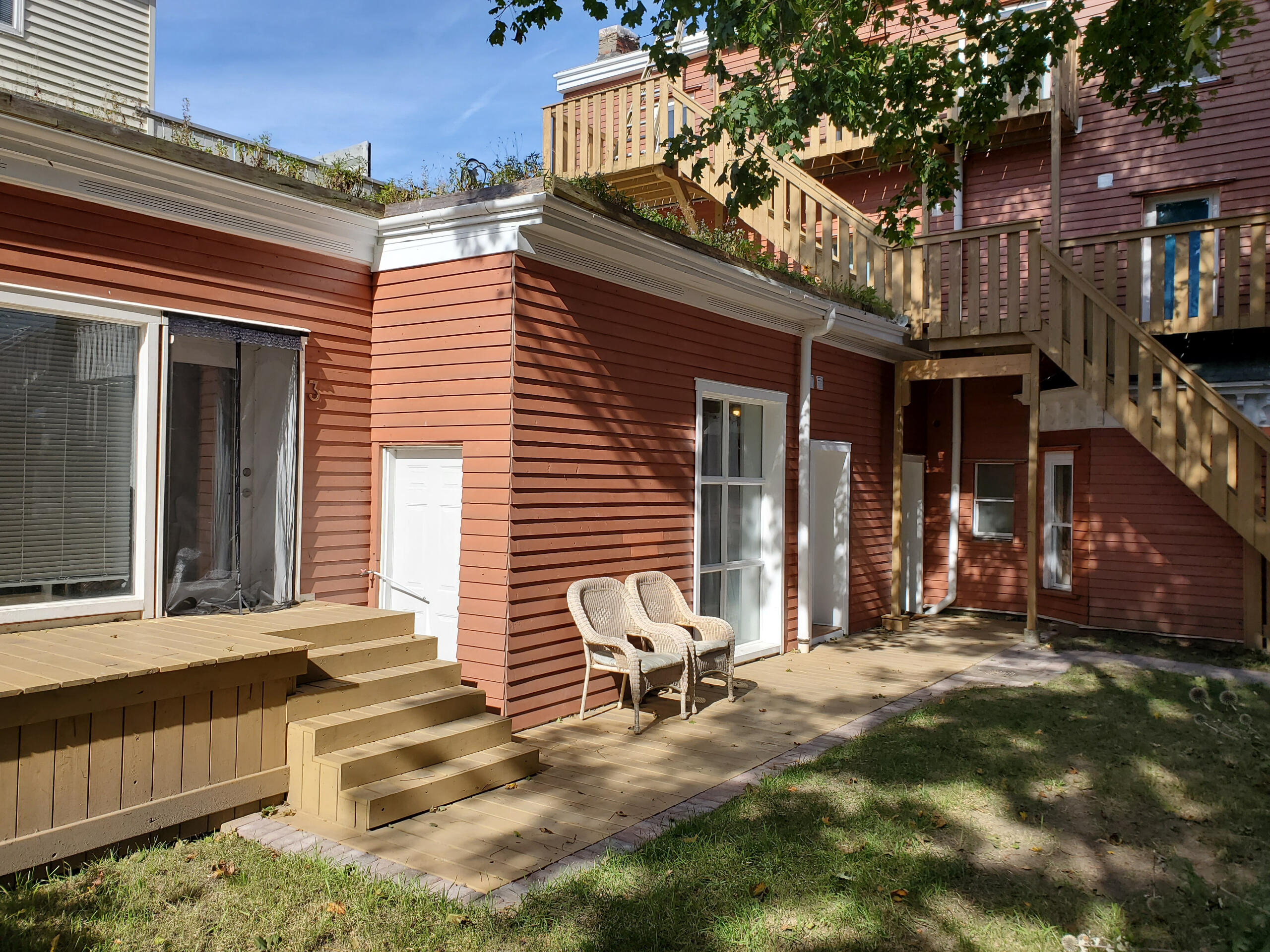 Image of the rear backyard of a home we worked on. Bright summer day with dappled sunlight through a tree. Light brick-colour stained house with trimmed lawn and two wicker chairs on a floor-level deck that has one small staircase to the left.